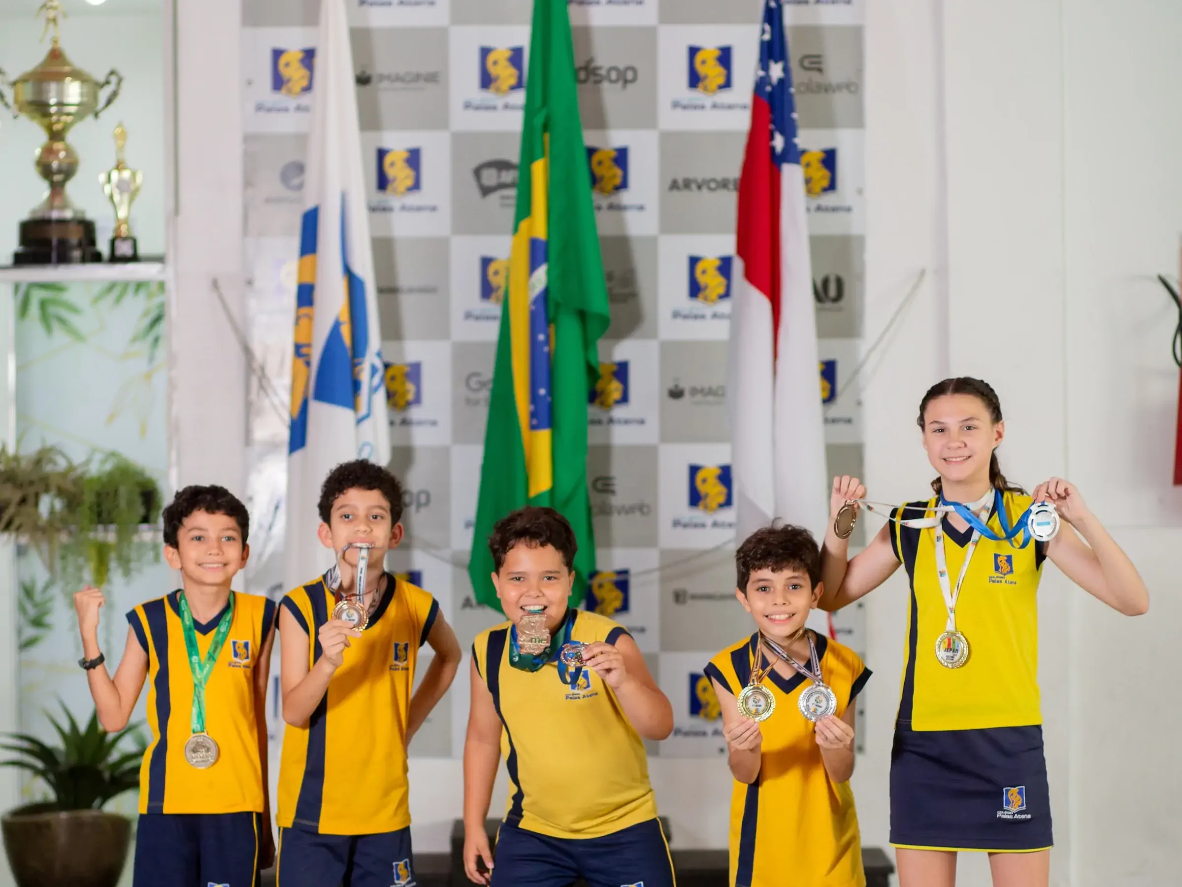 Alunos do Palas Atena com medalhas, diante das bandeiras e dos troféus da escola